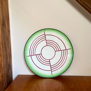 Puede incluir: Un plato de cerámica blanco con un borde verde y un diseño geométrico rojo. El diseño presenta círculos concéntricos y líneas.