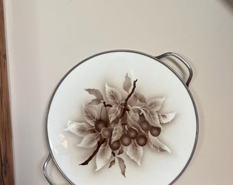 Op de afbeelding: Een witte keramische serveerschaal met een zilveren rand en handvatten. De schaal heeft een bruine en witte illustratie van een kersenboomtak met bladeren en vruchten.