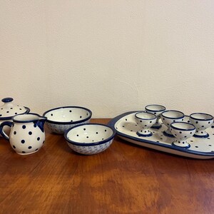 Può includere: Una collezione di stoviglie in ceramica bianca con pois blu. Include un piatto con coperchio, una caraffa per panna, due ciotole e un vassoio da portata con sei portauova. Il set è disposto su una superficie di legno.