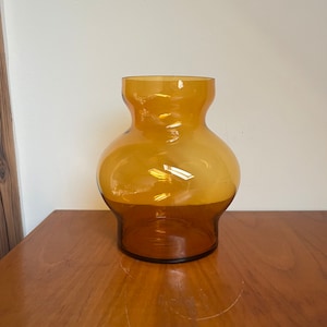 May include: A amber glass vase with a wide, rounded body and a narrow neck. The vase is sitting on a wooden surface.