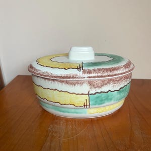 May include: A white ceramic bowl with a lid. The bowl has a brown, yellow, and green geometric pattern. The lid has a white knob.
