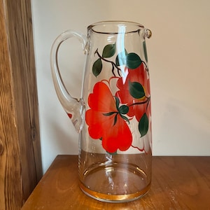 Puede incluir: Una jarra de vidrio transparente con un borde dorado y un diseño floral. Las flores son rojas, blancas y verdes.