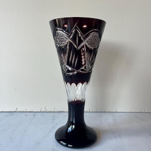May include: A tall, ruby-colored crystal glass with intricate etched designs. The glass features a flared top, a slender stem, and a rounded base. The etched patterns include geometric shapes and delicate lines, creating a sophisticated look.