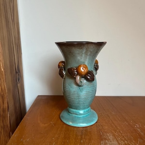 Può includere: Un vaso in ceramica turchese con accenti marroni e arancioni. Il vaso ha un corpo arrotondato e una parte superiore svasata. Presenta tre boccioli di fiori decorativi marroni e arancioni sul lato.