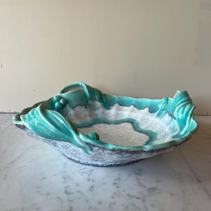 Op de afbeelding: Een keramische kom met een unieke, organische vorm. De kom heeft een turkooizen rand en accenten, met een wit en grijs interieur. De rand heeft een bladachtig ontwerp. De kom is waarschijnlijk bedoeld voor decoratieve doeleinden of om te serveren.