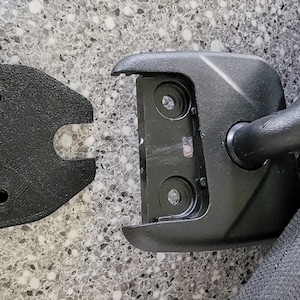 May include: Close-up of a black plastic car seat headrest hook with a removable cover. The hook is attached to a black fabric car seat.