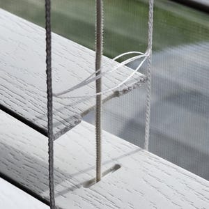 May include: Close-up of white wooden blinds with a textured surface. The blinds are partially open, revealing a glimpse of the outside. Vertical cords and horizontal slats create a pattern. The blinds are a neutral color.