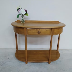 May include: A light brown, semi-circular console table with a single drawer and a lower shelf. The table has four slender legs and a small vase of flowers on top. The table is made of wood and has a smooth finish.
