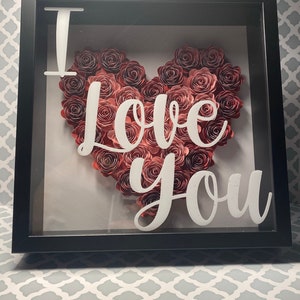 May include: A black shadow box with a white "I love you" inscription and a heart-shaped arrangement of paper roses in shades of pink and red.