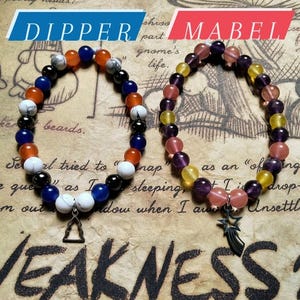 May include: Two beaded bracelets with charms. The bracelet on the left is blue, orange, white, and black with a triangle charm. The bracelet on the right is purple, pink, yellow, and orange with a star charm. The bracelets are on a brown background with the words "DIPPER" and "MABEL" above them.