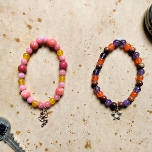 May include: Two beaded bracelets with charms. The first bracelet has pink, yellow, and orange beads with a rose charm. The second bracelet has purple, orange, and brown beads with a star charm.