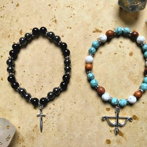 May include: Two beaded bracelets with charms. The first bracelet is black with a silver sword charm. The second bracelet has turquoise, white, and brown beads with a silver bow and arrow charm.