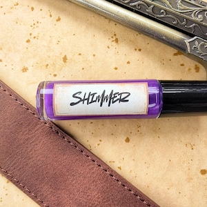 May include: A small purple bottle with a white label that says "SHIMMER" on a brown surface.