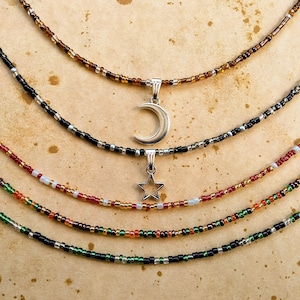 May include: Five layered beaded necklaces featuring small glass beads in various colors including brown, black, silver, red, green, and orange. Two of the necklaces feature silver-toned metal pendants, one shaped like a crescent moon and one like a star.