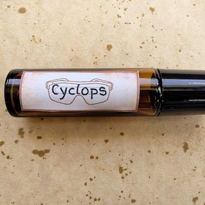 May include: A small amber glass roller bottle with a black cap and a white rectangular label. The label features a hand-drawn illustration of safety goggles with the word Cyclops written in black ink across the center of the goggles.