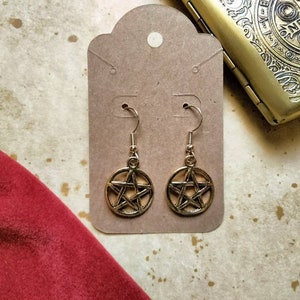 May include: A pair of gold-toned earrings with a pentagram design. The earrings are hanging from a card with a hole punched in the top.