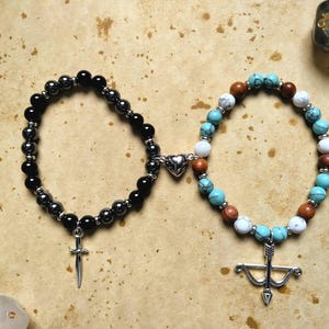 May include: Two beaded bracelets connected by a silver heart charm. One bracelet is black with a silver sword charm, the other is turquoise, white, and brown with a silver bow and arrow charm.