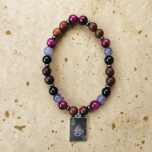 May include: A beaded bracelet with a silver charm featuring the Ace of Spades. The bracelet is made with brown, black, purple, and pink beads.