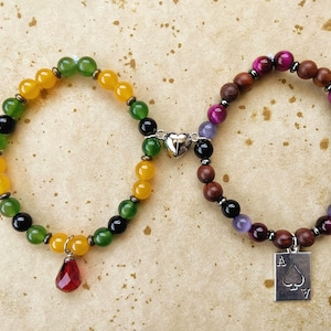 May include: Two beaded bracelets with charms. One bracelet has yellow, green, and black beads with a red teardrop charm. The other bracelet has brown, red, purple, and black beads with a silver charm shaped like the Ace of Spades.