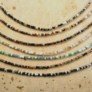 May include: A collection of seven layered necklaces made from small glass seed beads. The strands feature various color combinations including black, white, metallic gold, bronze, and green, arranged in repeating patterns along each individual string.