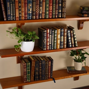K&ouml;nnte beinhalten: Drei Holzregale mit B&uuml;chern und Topfpflanzen. Das obere Regal hat eine Sammlung von ledergebundenen B&uuml;chern mit goldenen Beschriftungen. Das mittlere Regal hat eine Sammlung von B&uuml;chern mit goldenen und roten R&uuml;ckenteilen. Das untere Regal hat eine Sammlung von B&uuml;chern mit goldenen und blauen R&uuml;ckenteilen. Es gibt zwei Topfpflanzen, eine auf dem mittleren Regal und eine auf dem unteren Regal.