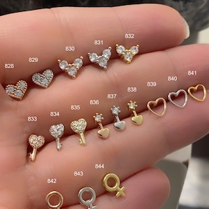 Peut inclure: Une variété de petites boucles d'oreilles en argent et en or avec différents motifs, notamment des cœurs, des clés et un symbole féminin. Les boucles d'oreilles sont numérotées de 828 à 844.
