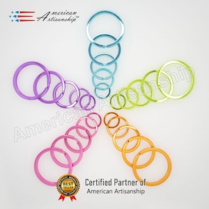 May include: Assortment of colorful metal key rings arranged in a starburst pattern. The key rings are in various sizes and colors, including purple, pink, orange, yellow, green, blue, and turquoise. The text "American Artisanship" is visible in the image.