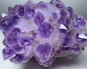 Può includere: Un grappolo di cristalli di ametista grezzi con punte viola intenso e violetto traslucido. La formazione minerale presenta una superficie frastagliata e sfaccettata con varie tonalità di viola, dal lilla chiaro al viola scuro intenso.
