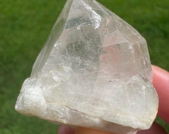 Puede incluir: Un cristal de cuarzo transparente con una forma geométrica puntiaguda. El cristal es translúcido con una apariencia ligeramente nublada y una base rugosa y texturizada. El fondo es verde borroso, lo que sugiere un entorno al aire libre. El cristal se sostiene en una mano.