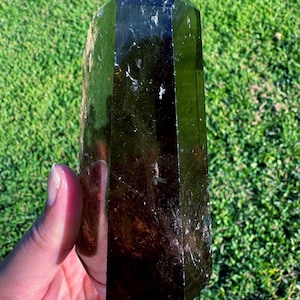 Può includere: Una torre di cristallo marrone scuro lucidata, con una parte superiore a punta. Il cristallo ha un design sfaccettato ed è tenuto contro uno sfondo di erba verde. La superficie del cristallo riflette la luce, mostrando il suo colore intenso e la sua chiarezza.