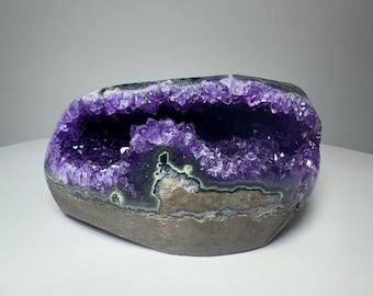 Può includere: Una grande geode di ametista caratterizzata da un interno cristallino viola intenso con formazioni di quarzo appuntite e frastagliate. L'esterno è composto da uno strato di roccia ruvida grigio-marrone con bande di depositi minerali verdi e bianchi.