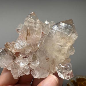 May include: A cluster of clear quartz crystals with golden-brown rutile inclusions. The crystals have sharp, pointed terminations and a translucent appearance, showcasing natural formations. The quartz is held in a hand.