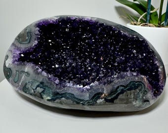 Può includere: Una grande geode di ametista ovale con un interno di cristalli viola scuro. L'esterno è una miscela di grigio, bianco e verde. Un oggetto decorativo naturale, perfetto per l'arredamento della casa.