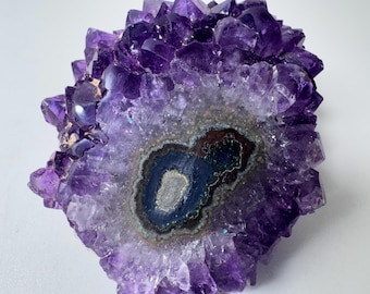 May include: A cluster of deep purple amethyst crystals featuring a central, dark, multi-layered agate formation with concentric rings of black, grey, and brown tones surrounded by jagged, translucent, and faceted crystalline points.