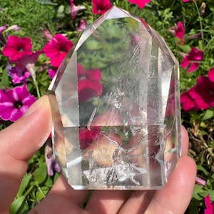 以下が含まれることがあります： 手で持たれた透明な水晶ポイント。 水晶は平らな上面と内部の亀裂が見えるようにカットされています。 背景には鮮やかなピンク色のペチュニアの花と緑の葉があり、自然な環境を作り出しています。