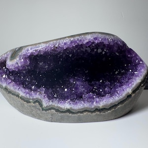 May include: A large, oval-shaped amethyst geode featuring deep purple, crystalline structures within a natural grey stone exterior. A black ruler is visible on the right side, showing a measurement of approximately 20 cm in length.