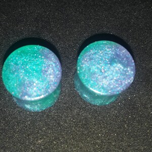 May include: Two ear plugs with a teal and purple glitter design. The plugs are round with a flat front and a slightly flared back. The glitter is embedded in a clear resin, creating a sparkling effect. The plugs are set against a black background.