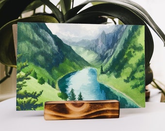 Pode incluir: Uma pintura em aquarela de uma cena de lago de montanha com um cavalete de madeira. A pintura apresenta um rio sinuoso, colinas verdes exuberantes e um céu nublado.