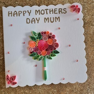 May include: A white Mother's Day card with a scalloped edge, featuring a paper flower bouquet and the words "HAPPY MOTHERS DAY MUM". The bouquet includes pink, orange, and green paper flowers. Small pink gems are scattered around the card.