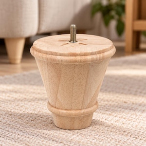 Puede incluir: Una pata de mueble de madera clara con forma cónica y estrecha, que presenta un anillo decorativo redondeado cerca de la base. Un perno roscado de metal sobresale de la parte superior circular plana, que tiene cuatro pequeñas muescas.