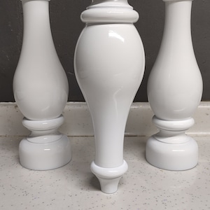 Puede incluir: Tres patas de mesa de madera blanca con un diseño torneado. Las patas son todas del mismo estilo, pero de diferentes tamaños. Las patas están sobre una superficie blanca con purpurina.