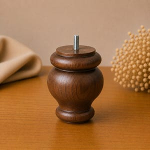 May include: A dark brown wooden furniture leg with a rounded, bulbous shape and a smooth, polished finish. The top of the leg features a centered, silver-colored metal threaded bolt protruding upwards for attachment to a furniture base.