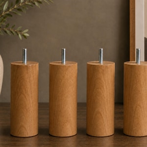 May include: Four cylindrical furniture legs made of light brown wood with a visible natural grain pattern. Each leg features a vertical silver metal threaded bolt protruding from the centre of the flat top surface for attachment purposes.