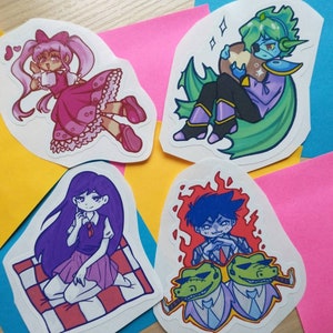 Könnte beinhalten: Vier bunte Aufkleber mit Comicfiguren. Der erste Aufkleber zeigt ein Mädchen mit rosa Haaren in einem rosa Kleid, das eine Tasse hält. Der zweite Aufkleber zeigt eine grünhäutige Figur mit blauen Haaren, die ein braunes Objekt hält. Der dritte Aufkleber zeigt ein Mädchen mit lila Haaren in einem rosa und weißen Kleid, das auf einer karierten Decke sitzt. Der vierte Aufkleber zeigt eine Figur mit blauen Haaren vor einem roten Hintergrund mit zwei grünen Krokodilköpfen.