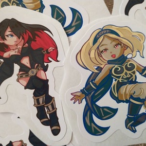 May include: Two cartoon stickers of female characters with black and gold outfits. The character on the left has long black hair with red highlights and a black cape. The character on the right has long blonde hair and a blue cape.