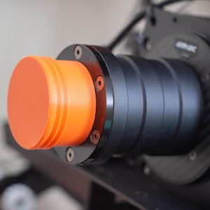 Puede incluir: Primer plano de un objetivo de cámara negro con una tapa naranja. El objetivo tiene una serie de anillos alrededor y la tapa tiene una superficie texturizada.