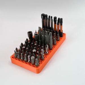 May include: An orange rectangular organizer filled with various dark gray and black screwdriver bits. The bits are of different shapes and sizes, including flathead, Phillips head, and Torx bits. The organizer is on a white background.