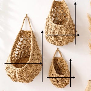 May include: Three woven jute hanging baskets of different sizes, suspended from brass hooks on a white wall. The baskets have open, textured weaves and a natural, light brown colour, suitable for storage or decoration.
