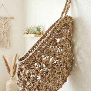 May include: A woven jute wall basket with a hanging loop, suspended from a gold hook on a white wall. The basket has an open weave design and is filled with air plants. A macrame wall hanging and vase are visible in the background.