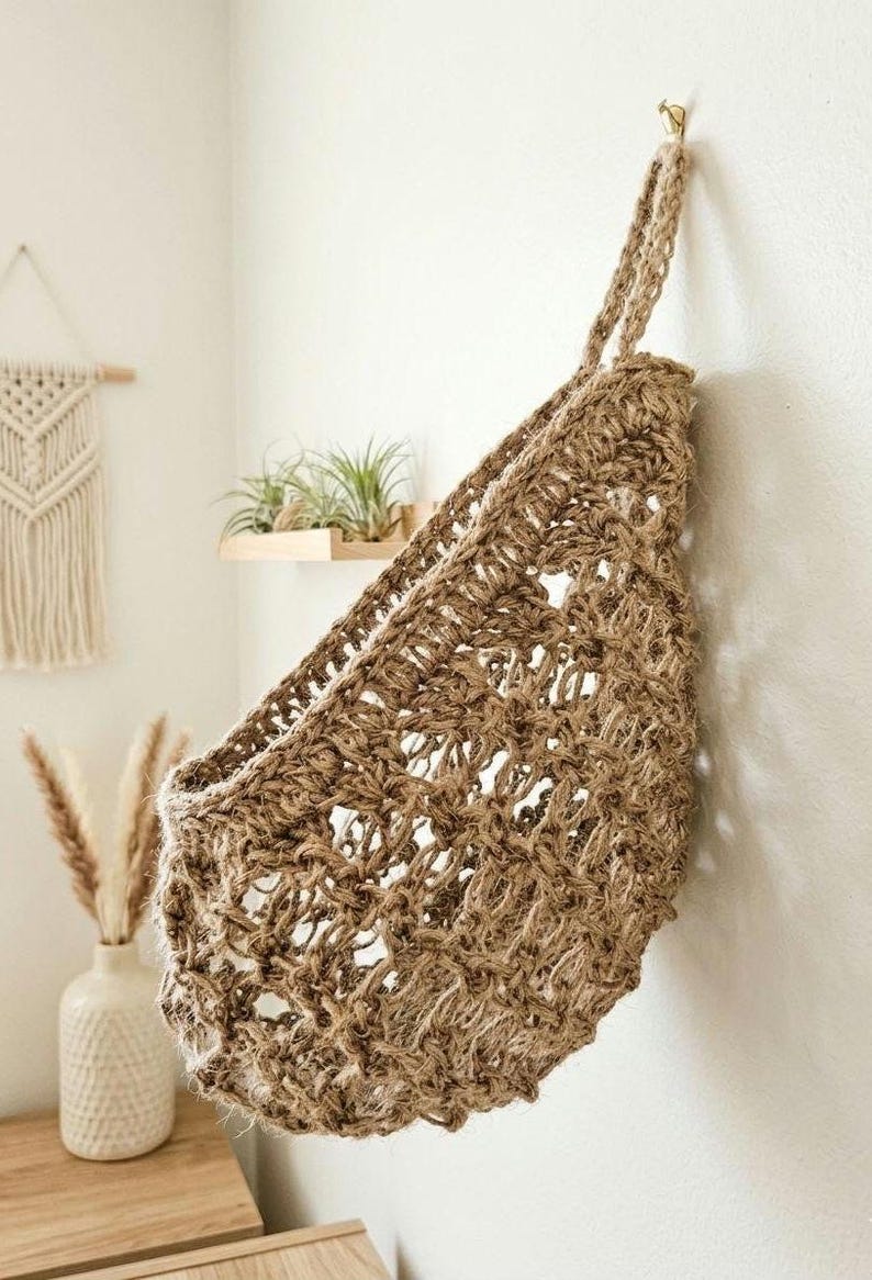 May include: A woven jute wall basket with a hanging loop, suspended from a gold hook on a white wall. The basket has an open weave design and is filled with air plants. A macrame wall hanging and vase are visible in the background.